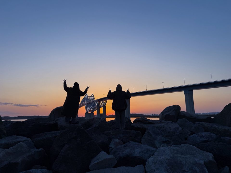 夕暮れの海辺の岩場で、橋を背にピースをする二人のシルエット