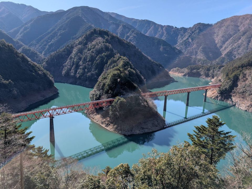 山に囲まれたエメラルド色の湖と、湖に突き出た岬をまたいで架かる赤い鉄橋